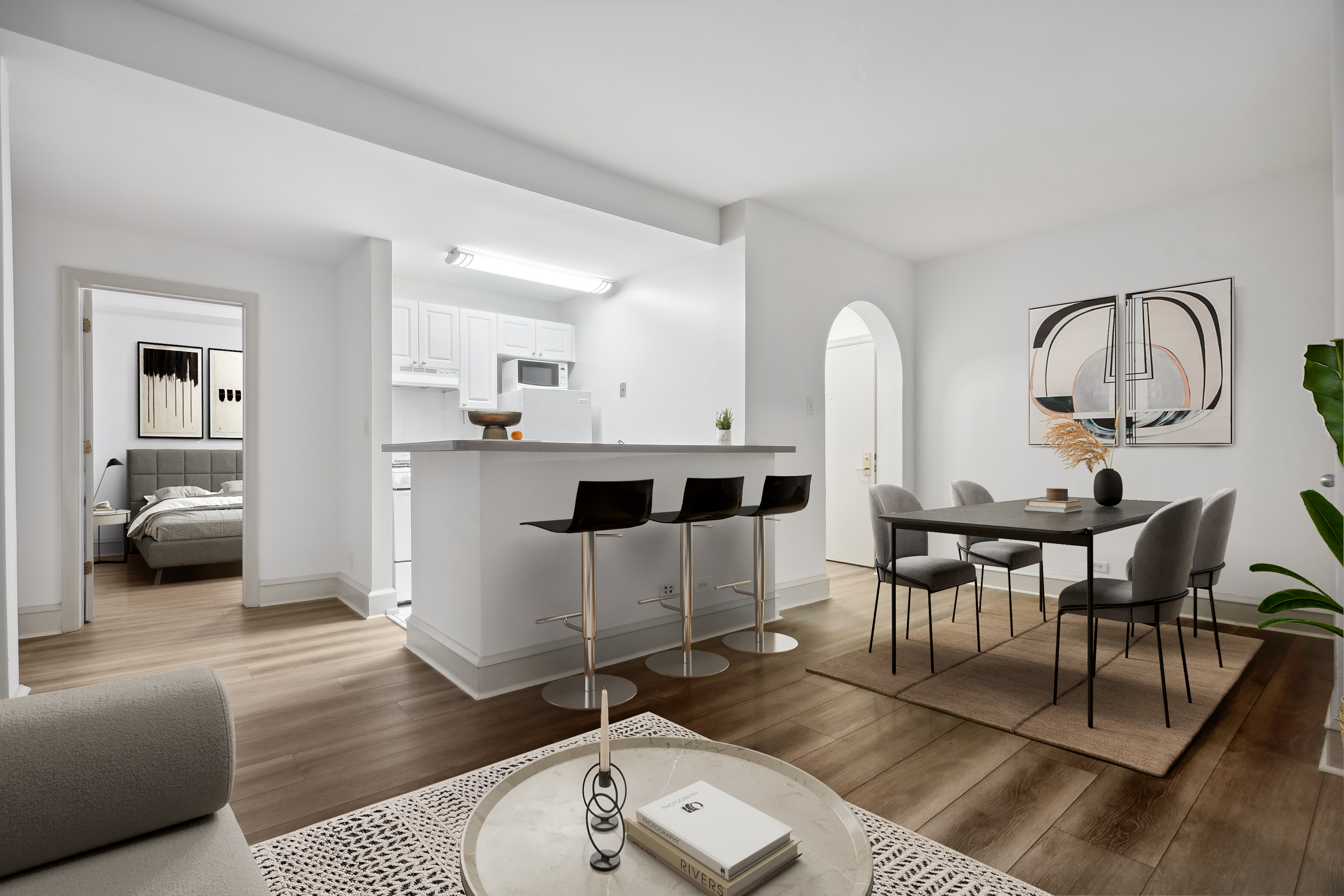 Renovated apartment with white cabinetry, kitchen island with seating for three.  View into bedroom on the left and dining table for 4 on the right.