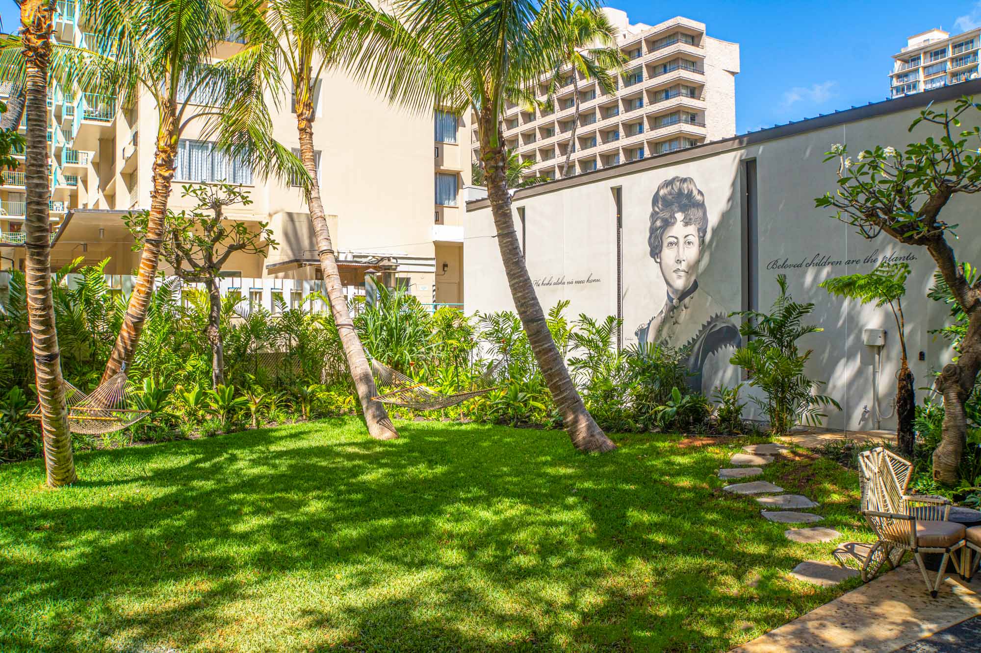 Grassy area at Lilia with hammocks hanging from palm trees and a mural of Queen Emma in the background.