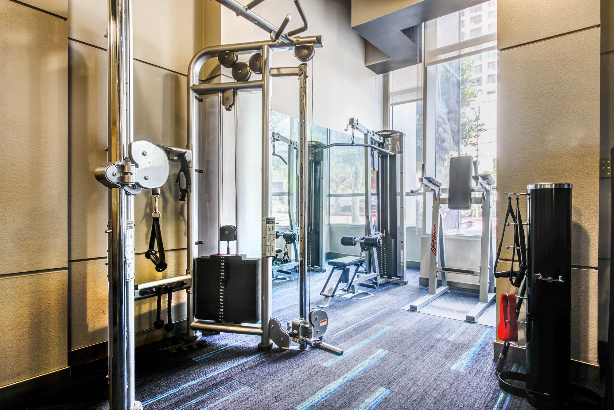 A fitness center area with cable machines by a window facing the street at The Met apartments in downtown LA