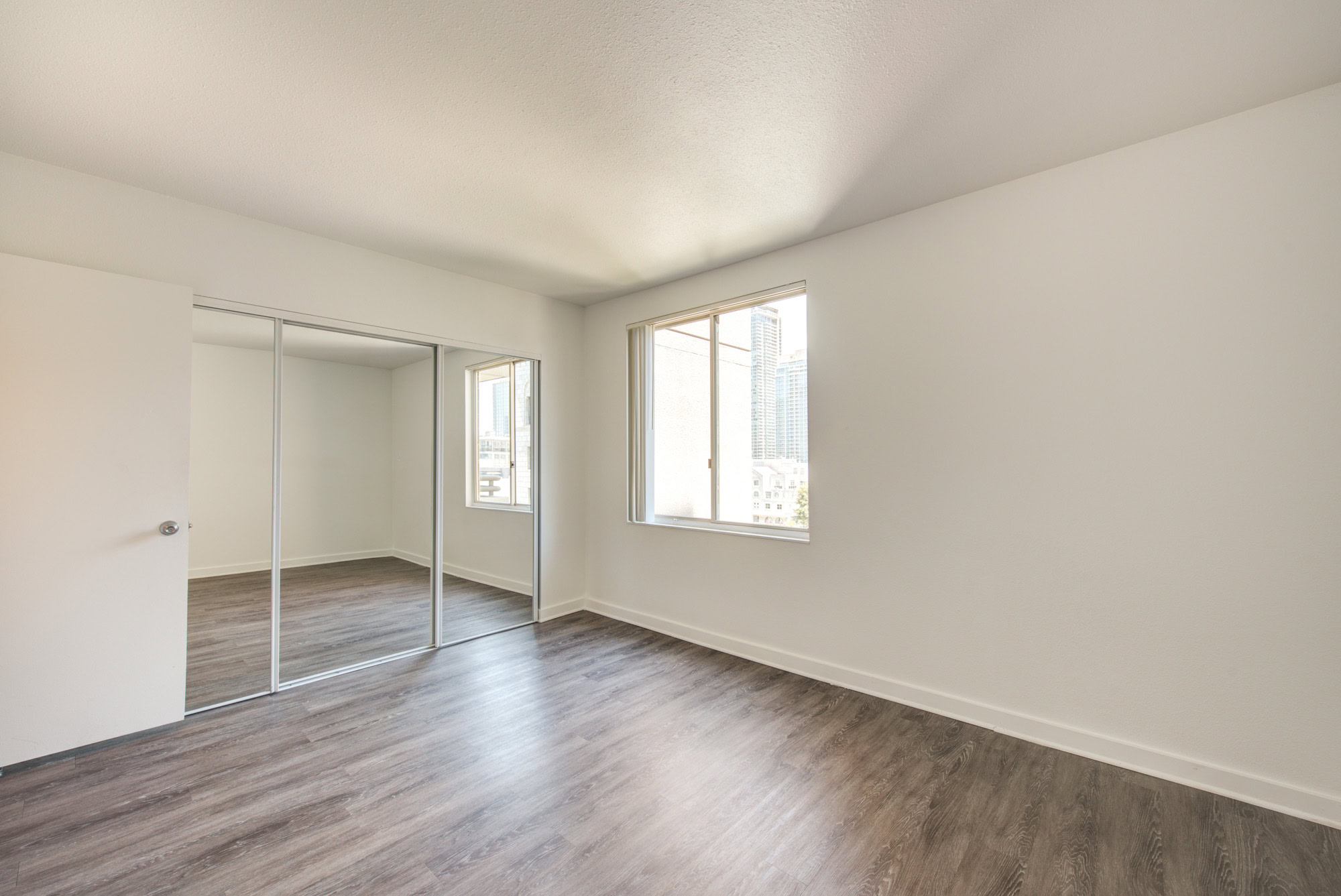 A bedroom with a closet with mirror doors, hard wood floors and a large window at The Mets modern apartments in downtown LA