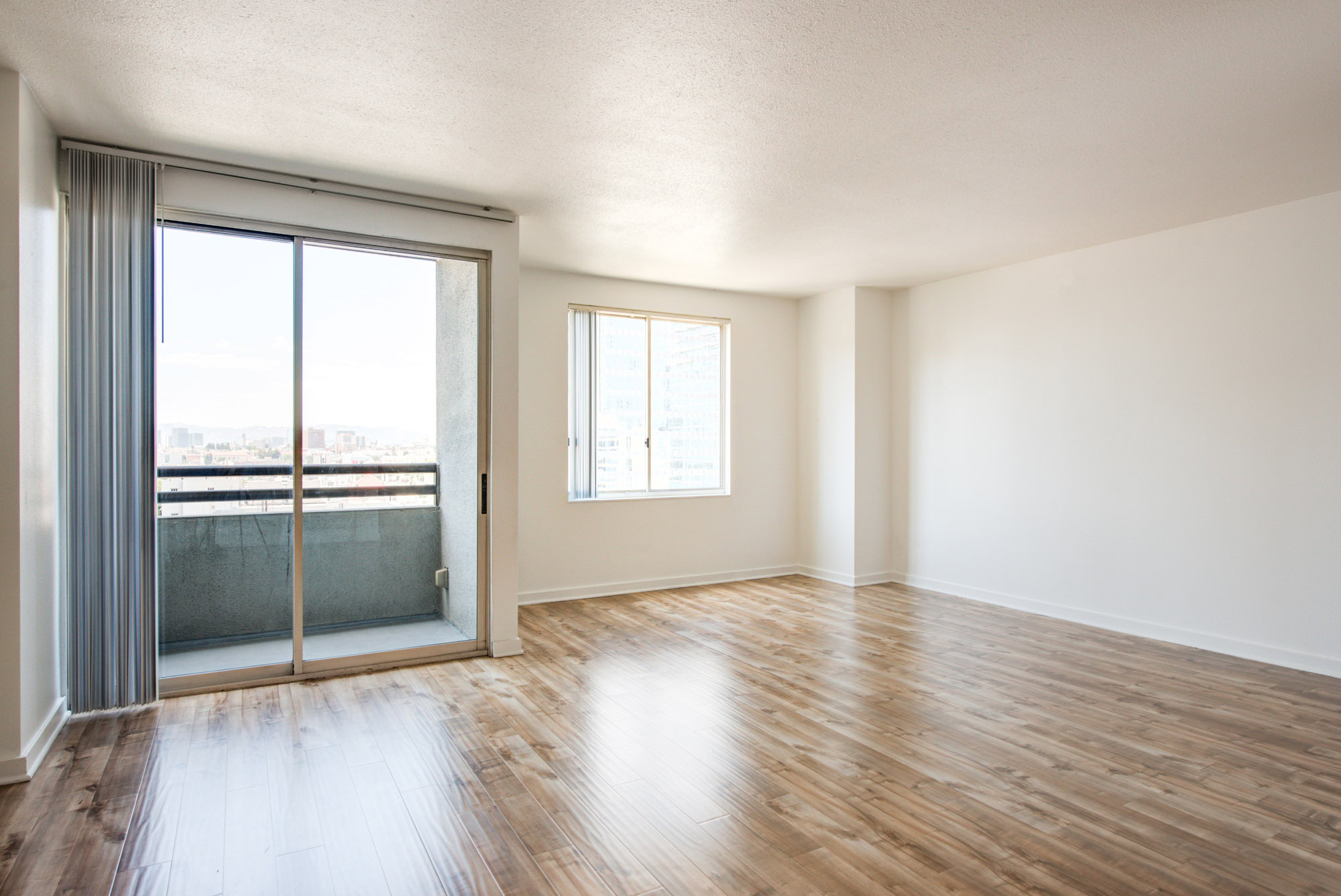 An empty studio living room with sliding doors to a balcony, a window and hardwood floors at The Mets modern apartments in Los Angeles