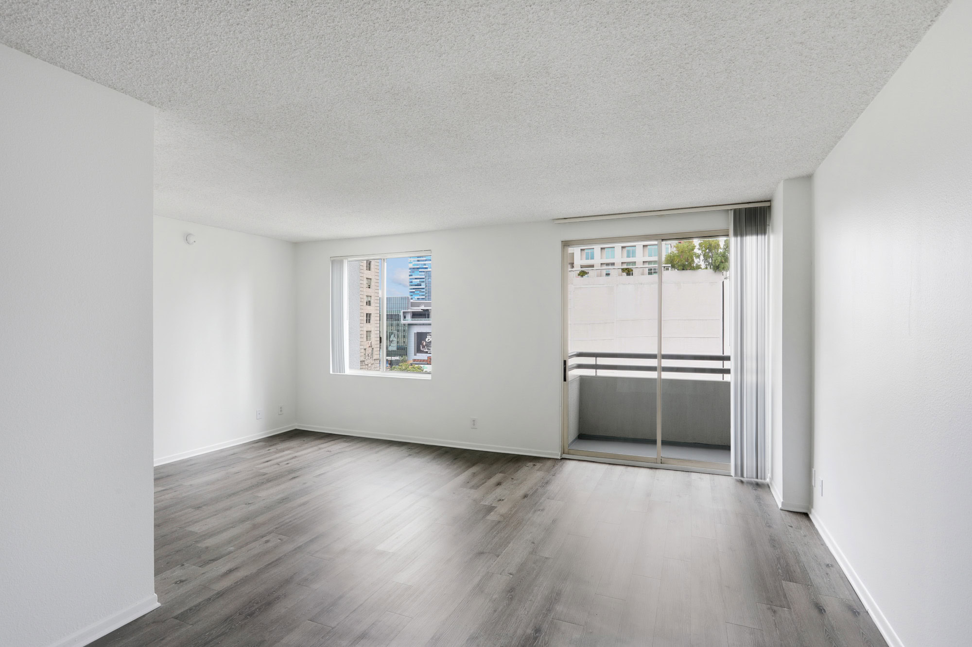 An empty studio living room with sliding doors to a balcony, a window and hardwood floors at The Mets modern apartments in Los Angeles