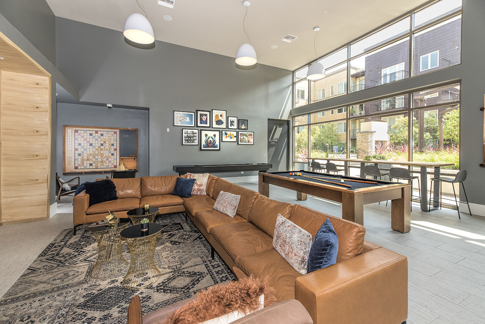 A game room with a large leather couch, a pool table and shuffle board with glass windows overlooking the courtyard at The Village Residences luxury apartments in Mountain View CA