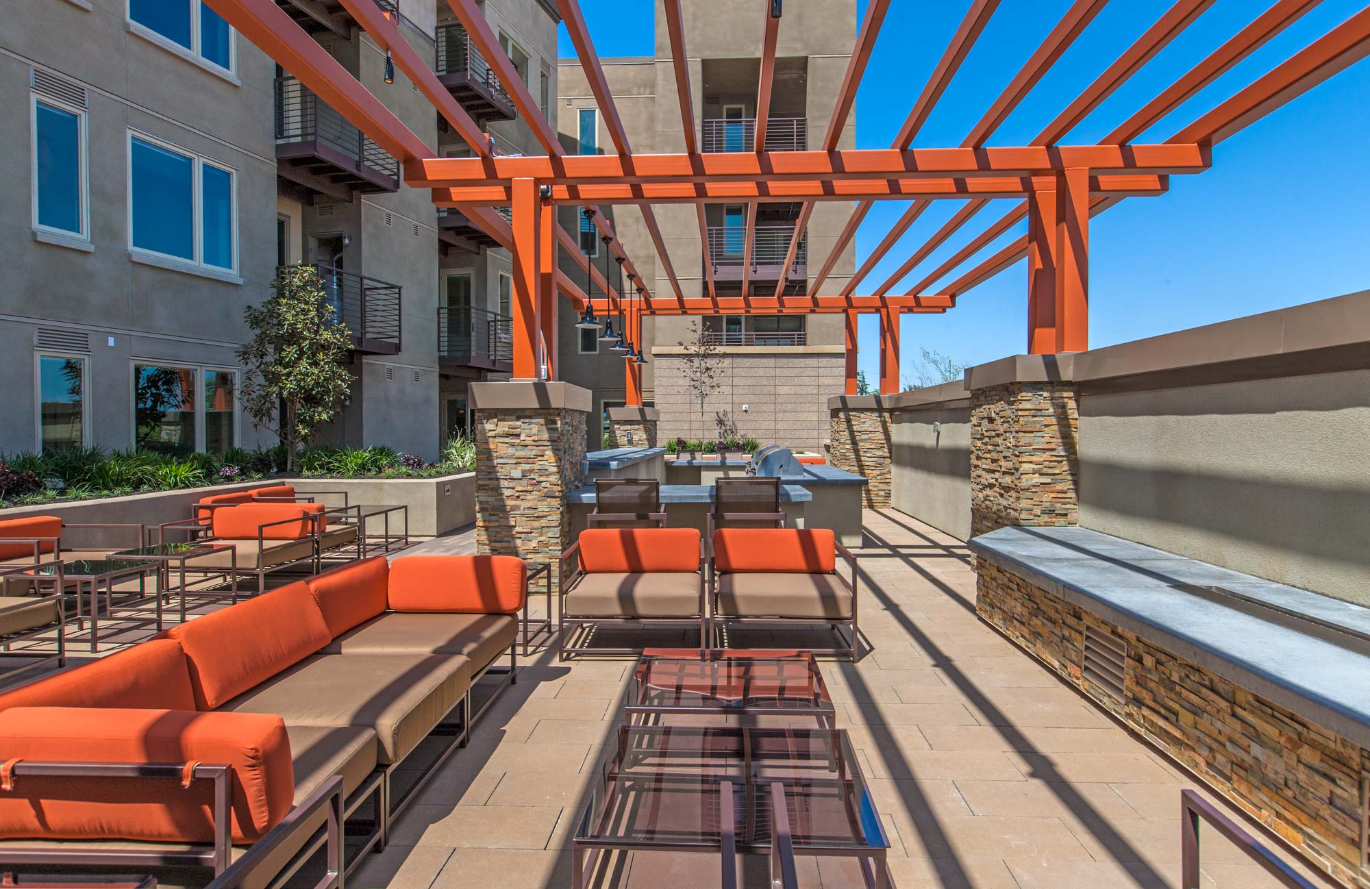 A patio with a several lounging areas, a grilling station and bar set up under an orange pergola at The Village Residences new apartments in Mountain View