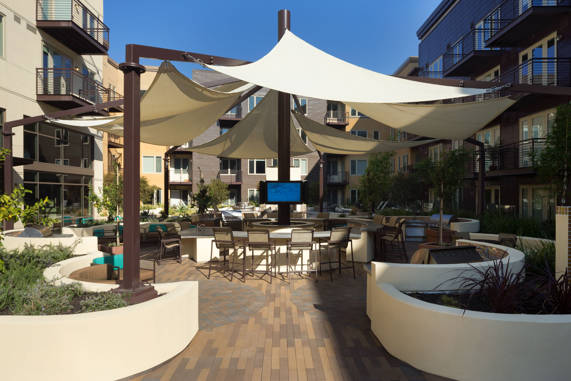 An outdoor courtyard entertaining area with several seasoning spaces, outdoor grilling stations, a bar and TV's at The Village Residences luxury apartments in Mountain View CA