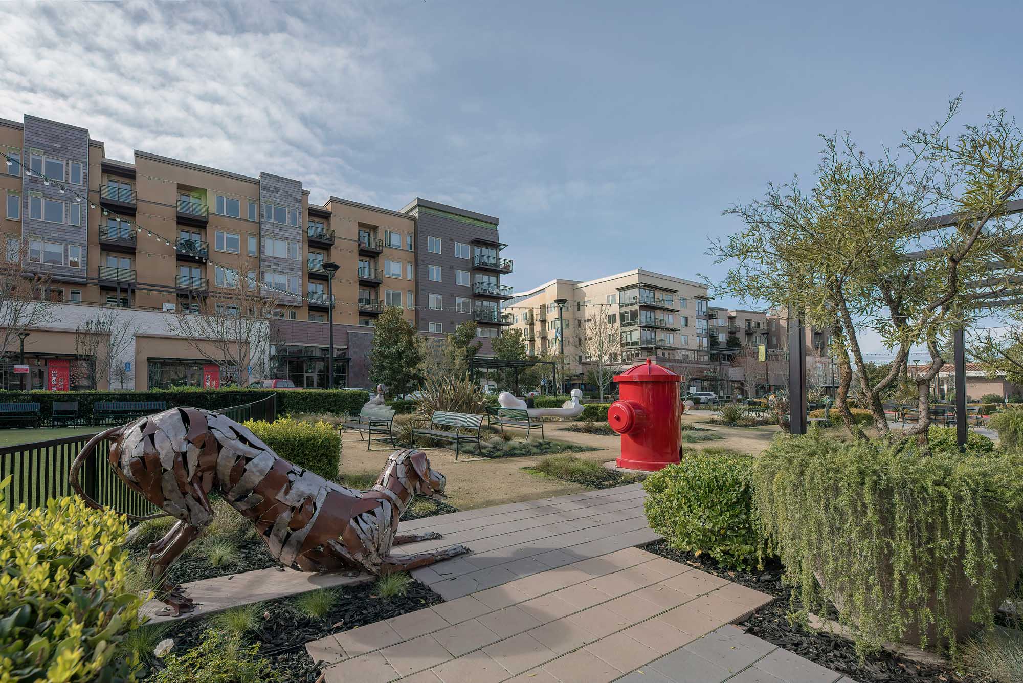 An outdoor courtyard with park benches and a gated dog yard featuring a giant red fire hydrant and a scrap metal dog sculpture at The Village Residences new apartments in Mountain View, CA