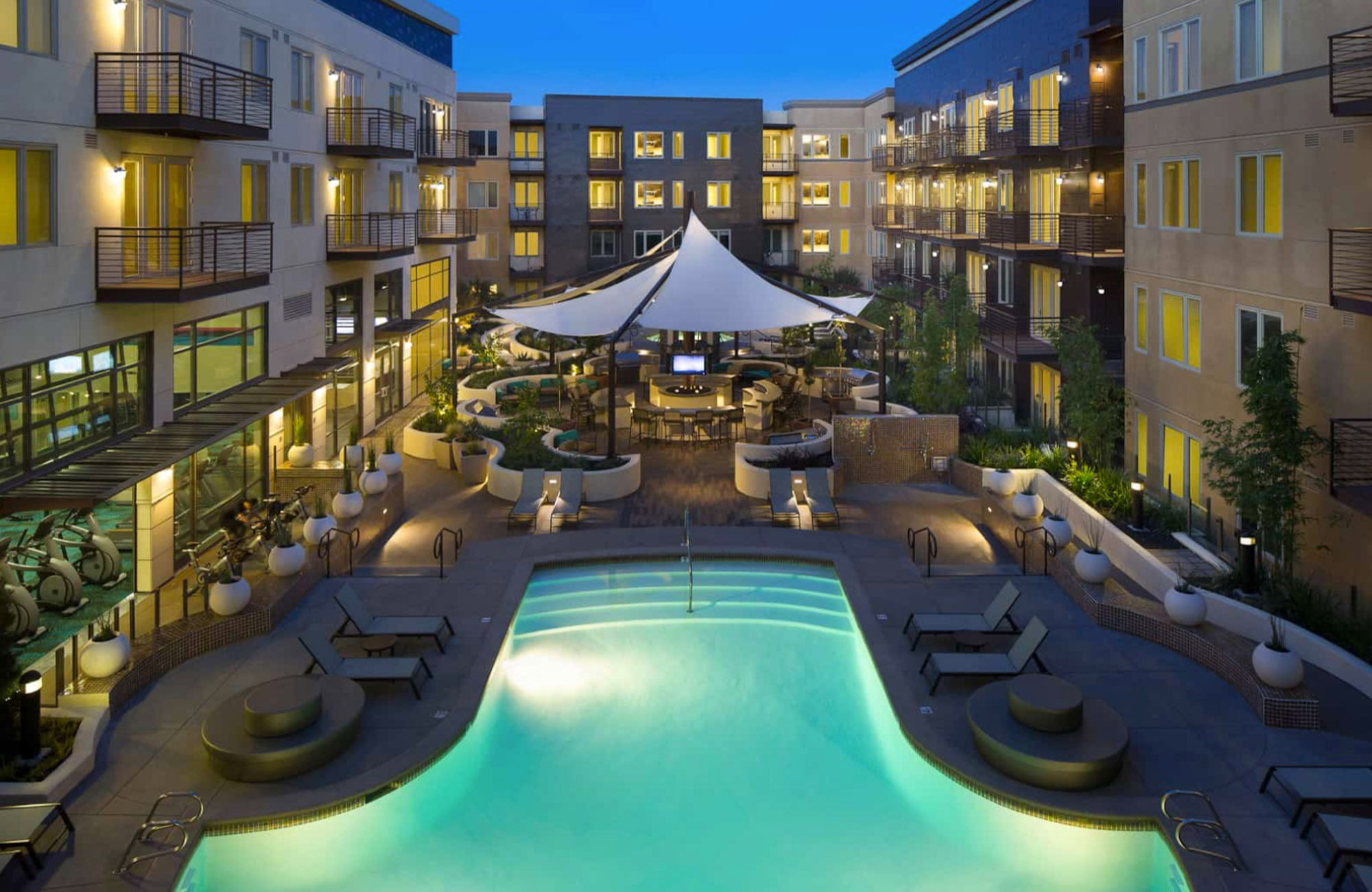 A spacious modern courtyard with a pool and lounging chairs, a bar area surrounding 360 tv's at The Village Residences Mountain View luxury apartments
