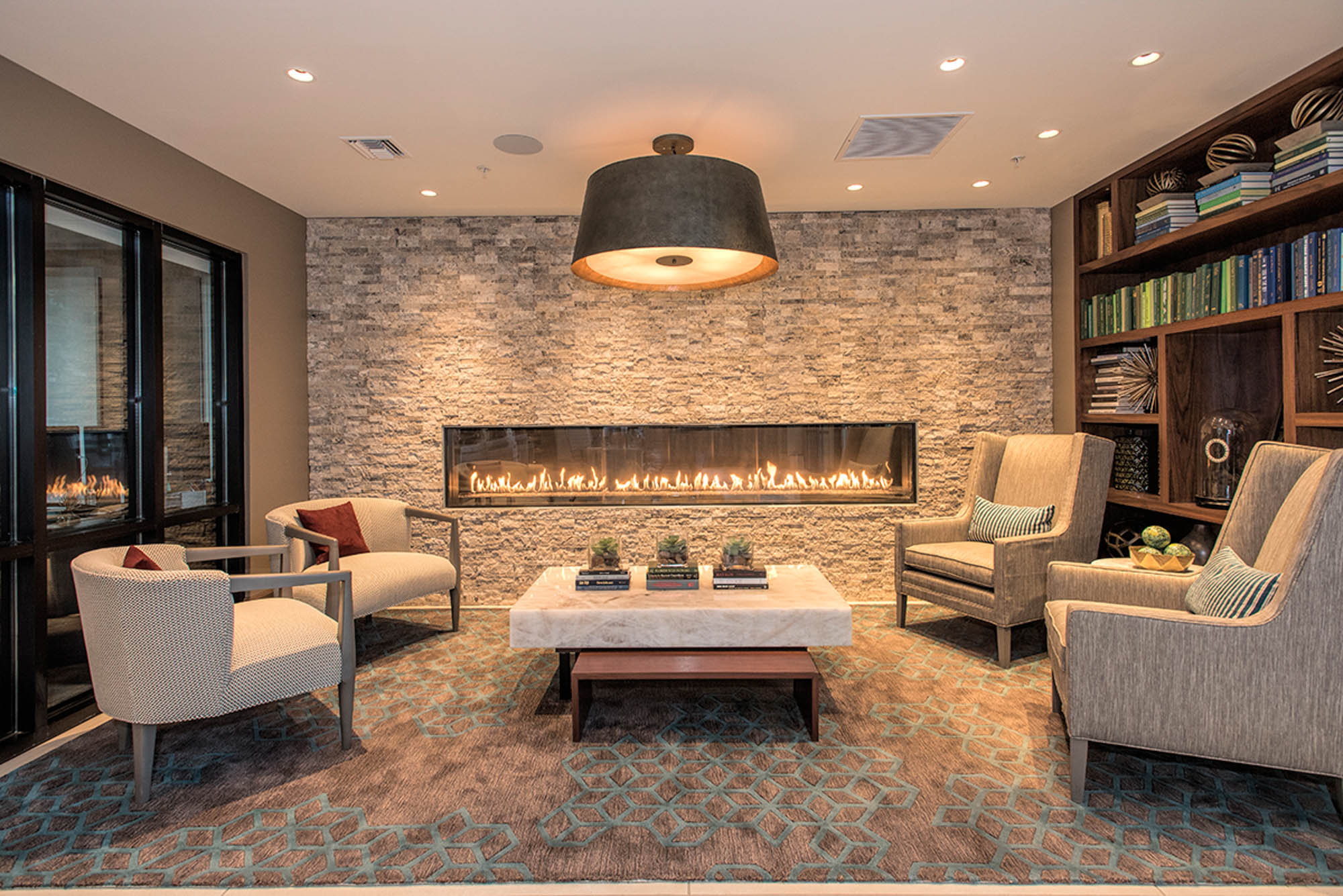 A lounge with a floor to ceiling library bookshelf, and a modern fireplace mounted on a stone accent wall at Vintage apartments in Pleasanton, CA