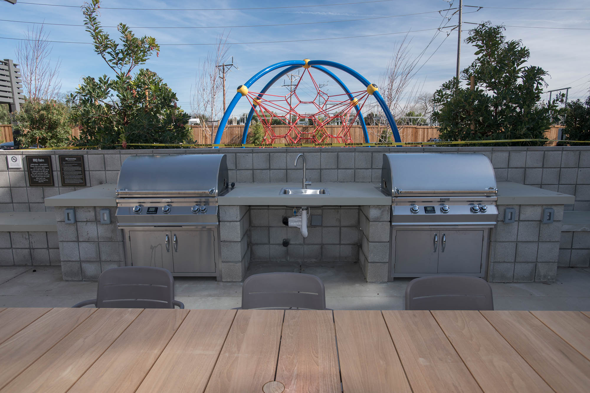 An outdoor grilling station with a dining table and a playground behind it, at the Vintage apartments in Pleasanton, CA