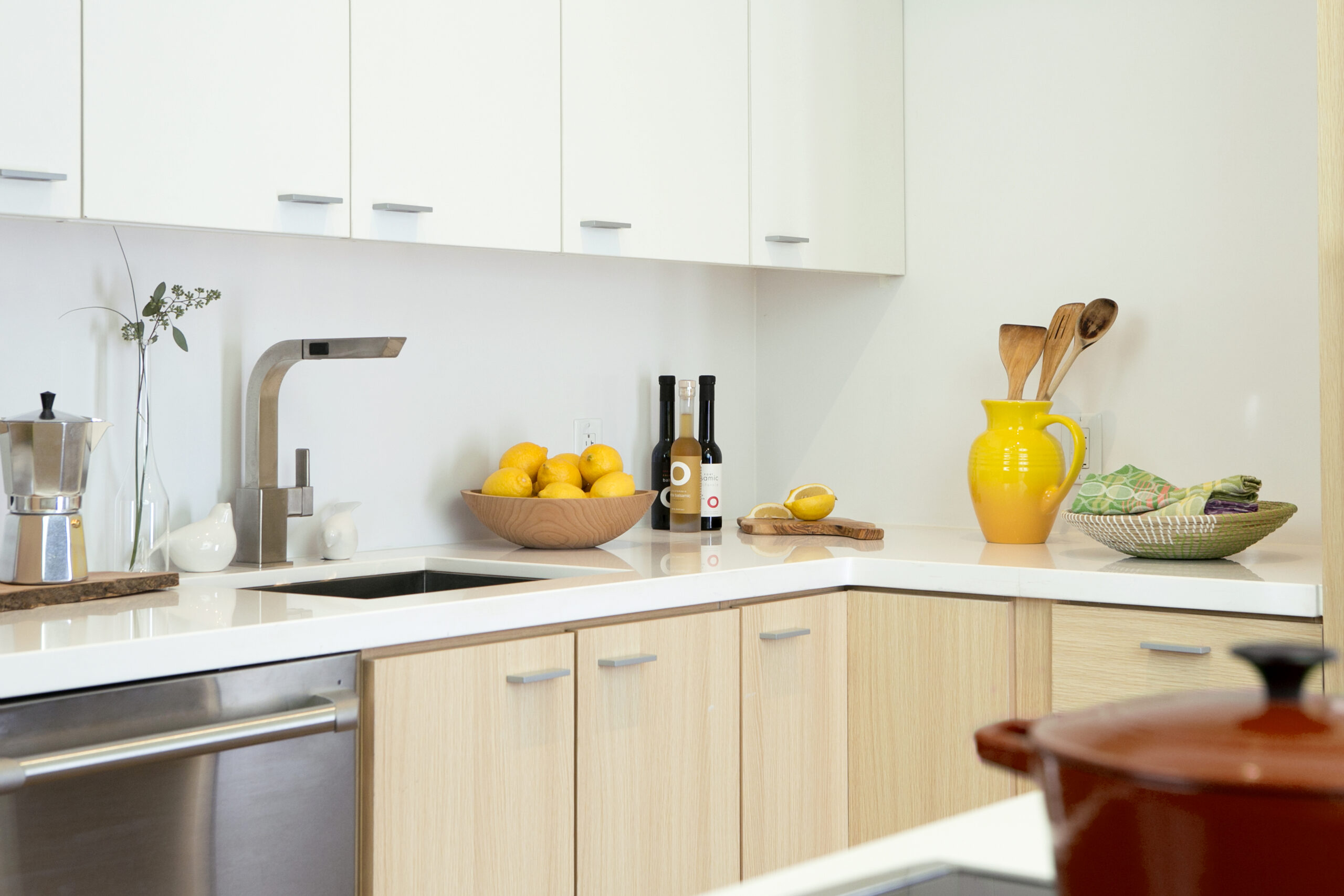 Bright kitchen with light wood cabinets, white countertops, and pops of yellow decor, including a pitcher and lemons in a bowl. Stainless steel appliances add a modern touch.