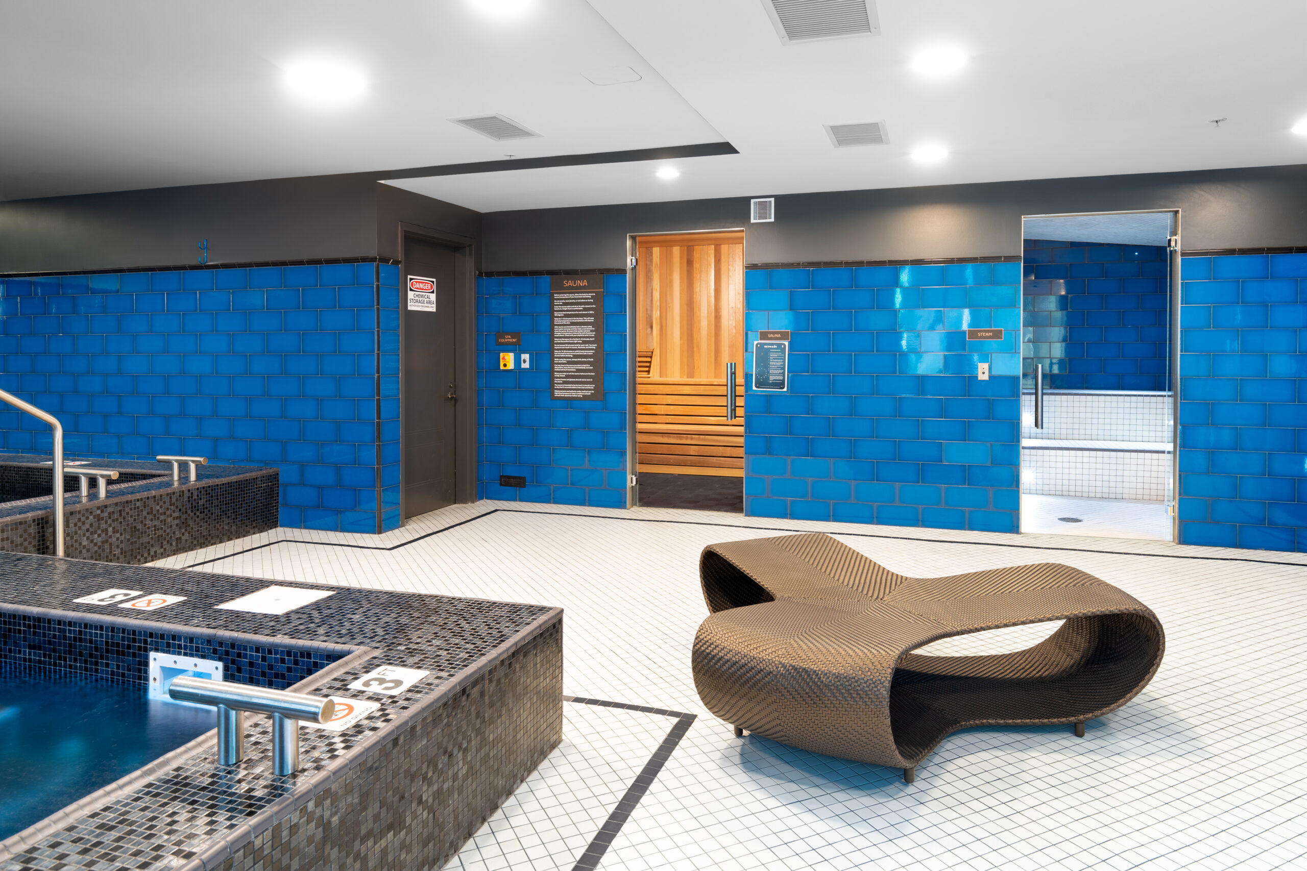 Spa area with blue tiled walls, sauna and steam room entrances, and a hot tub. Unique lounge chair in the center.