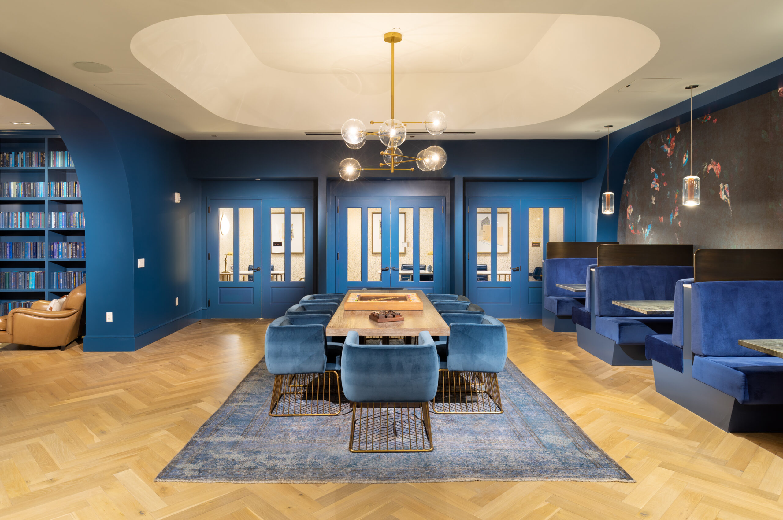 Elegant dining area with blue walls, wooden table, velvet chairs, and co-working stations.





