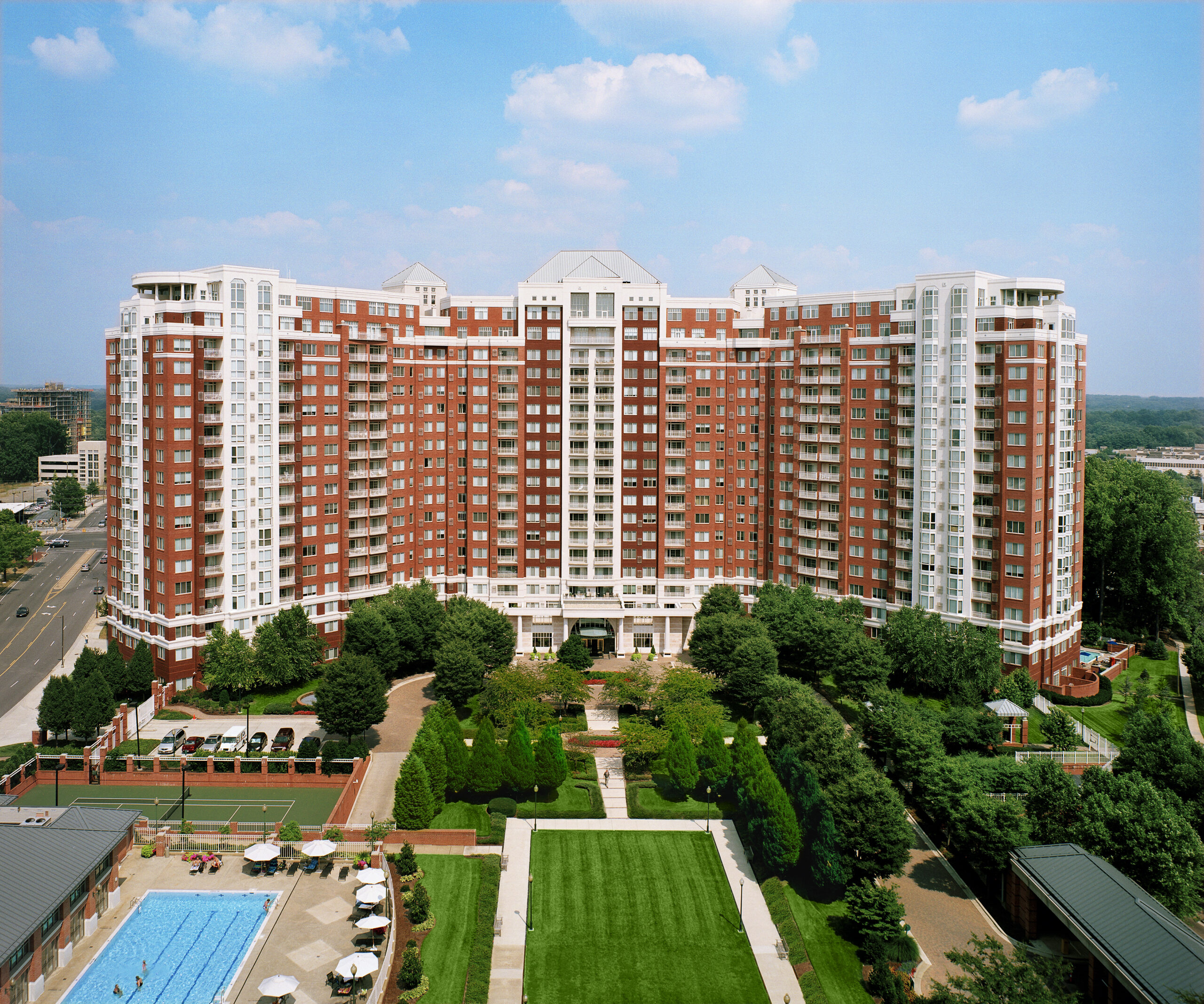 Large red-brick apartment complex with balconies, a landscaped garden, and a swimming pool, set against a blue sky.