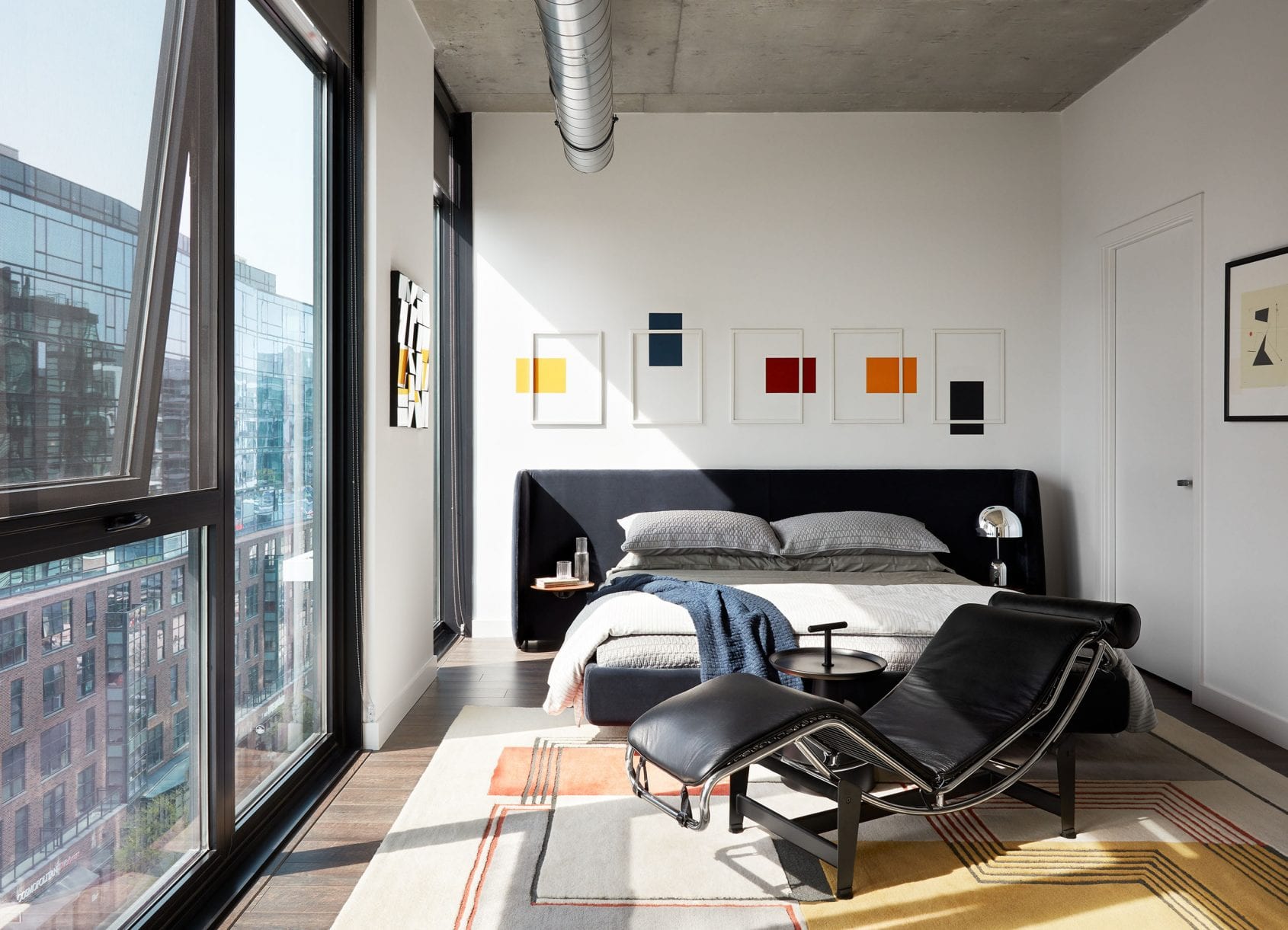 Bright bedroom with large windows, a black chaise lounge, and geometric wall art.