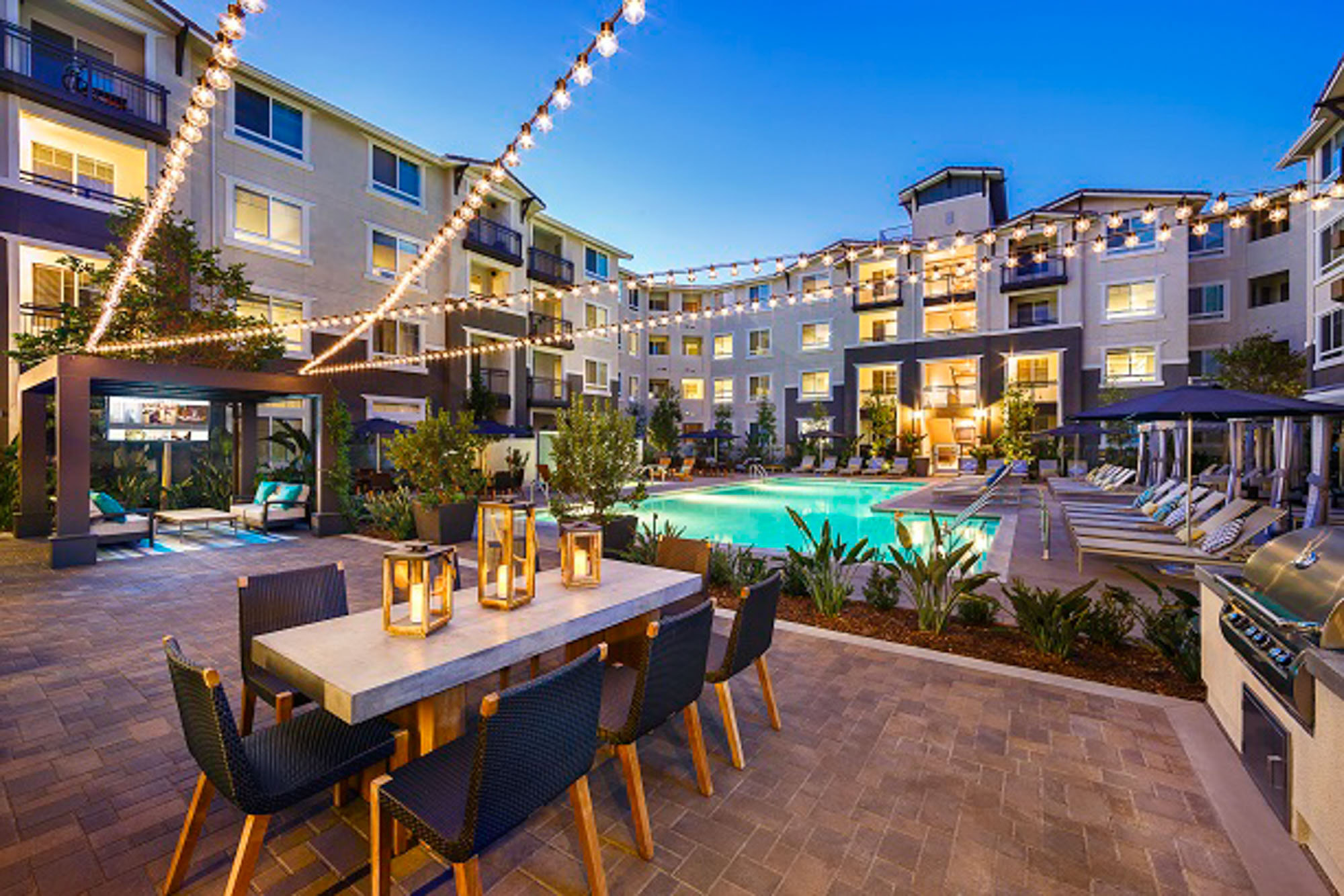 A beautifully lit outdoor patio an outdoor dining table, entertainment area, and lounging chairs next to be pool at the Adler luxury apartment in Northridge CA