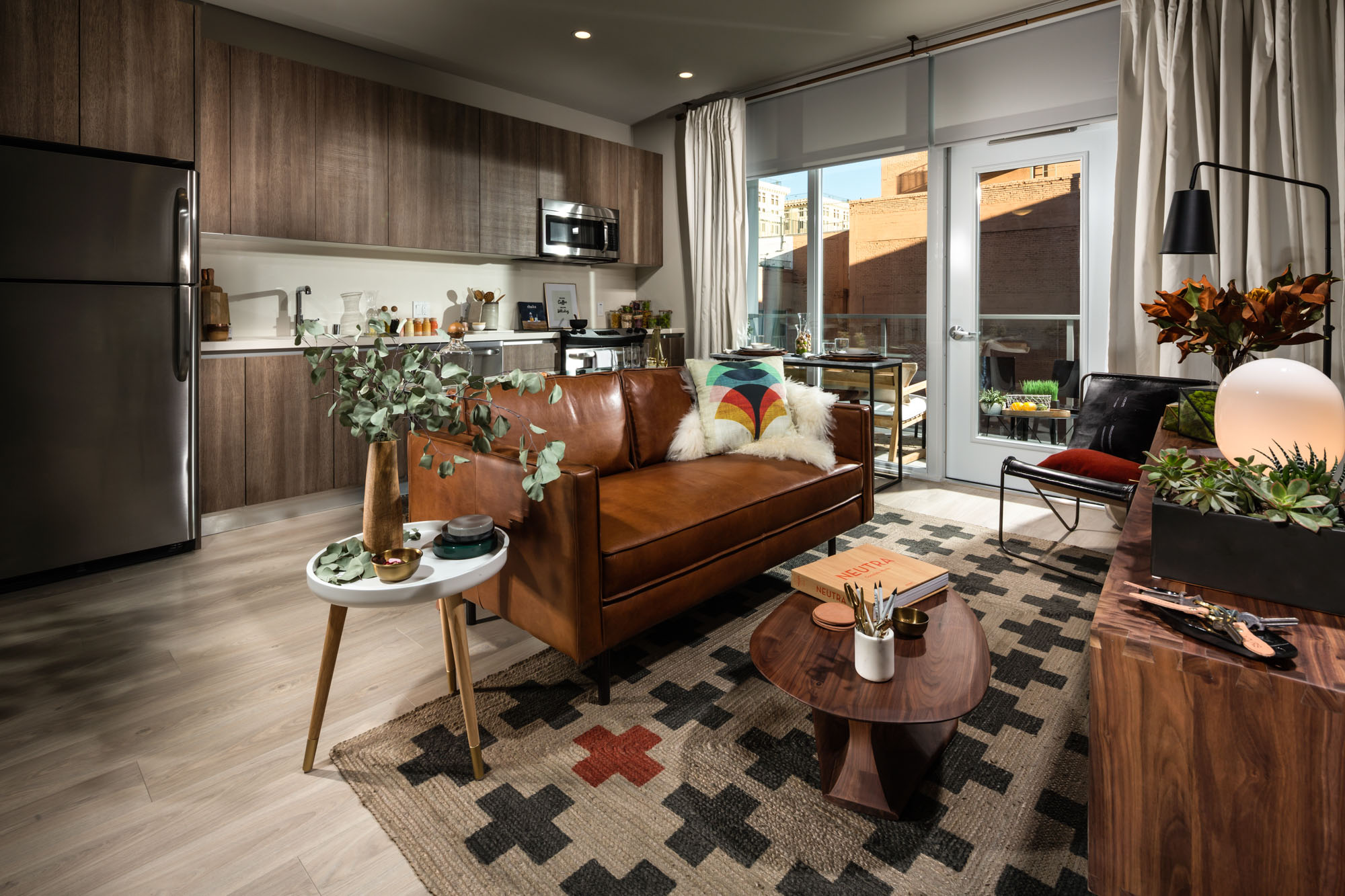 A living room and kitchen area with a glass door leading to a balcony, wood flooring and stainless steel kitchen appliances at 8th and Grand apartments in Downtown LA