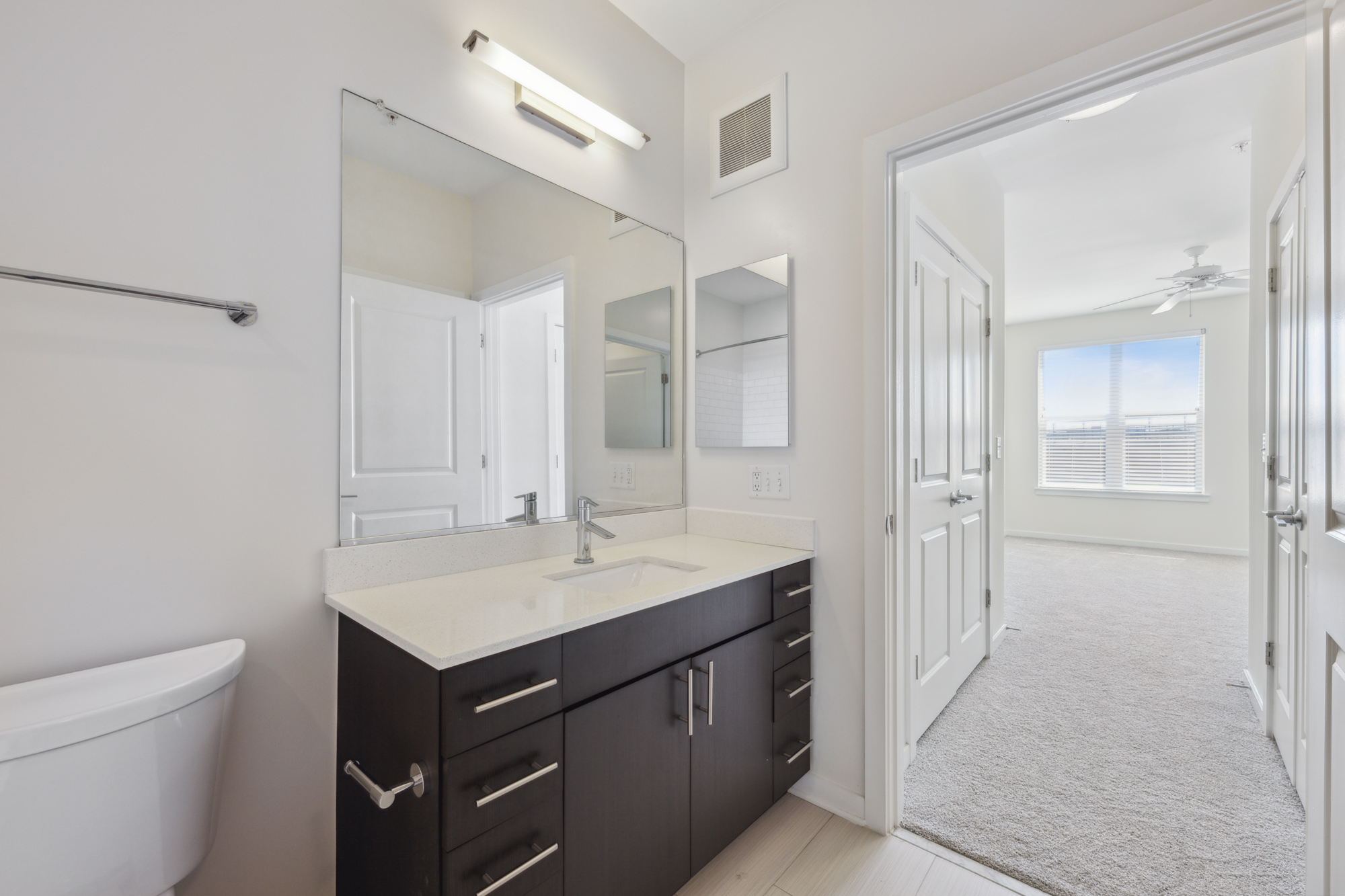 Bath with quartz countertops and dark cabinetry with bedroom entry in background.