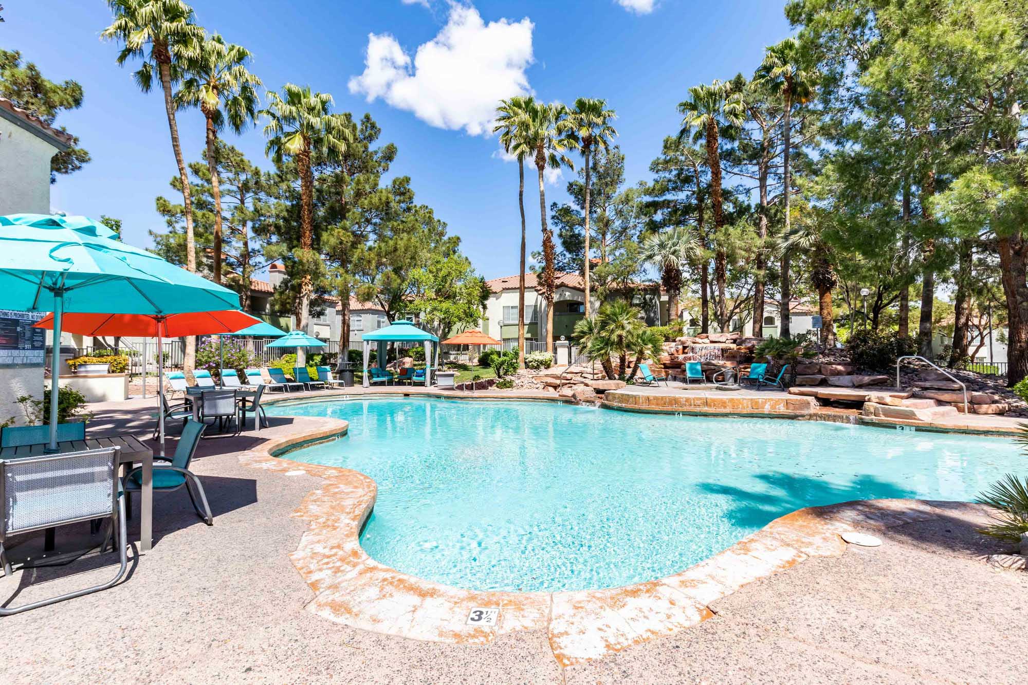 A resort-style, spanish style, pool area with palm trees with lounging chairs and umbrellas at Vintage apartments in Pleasanton, CA