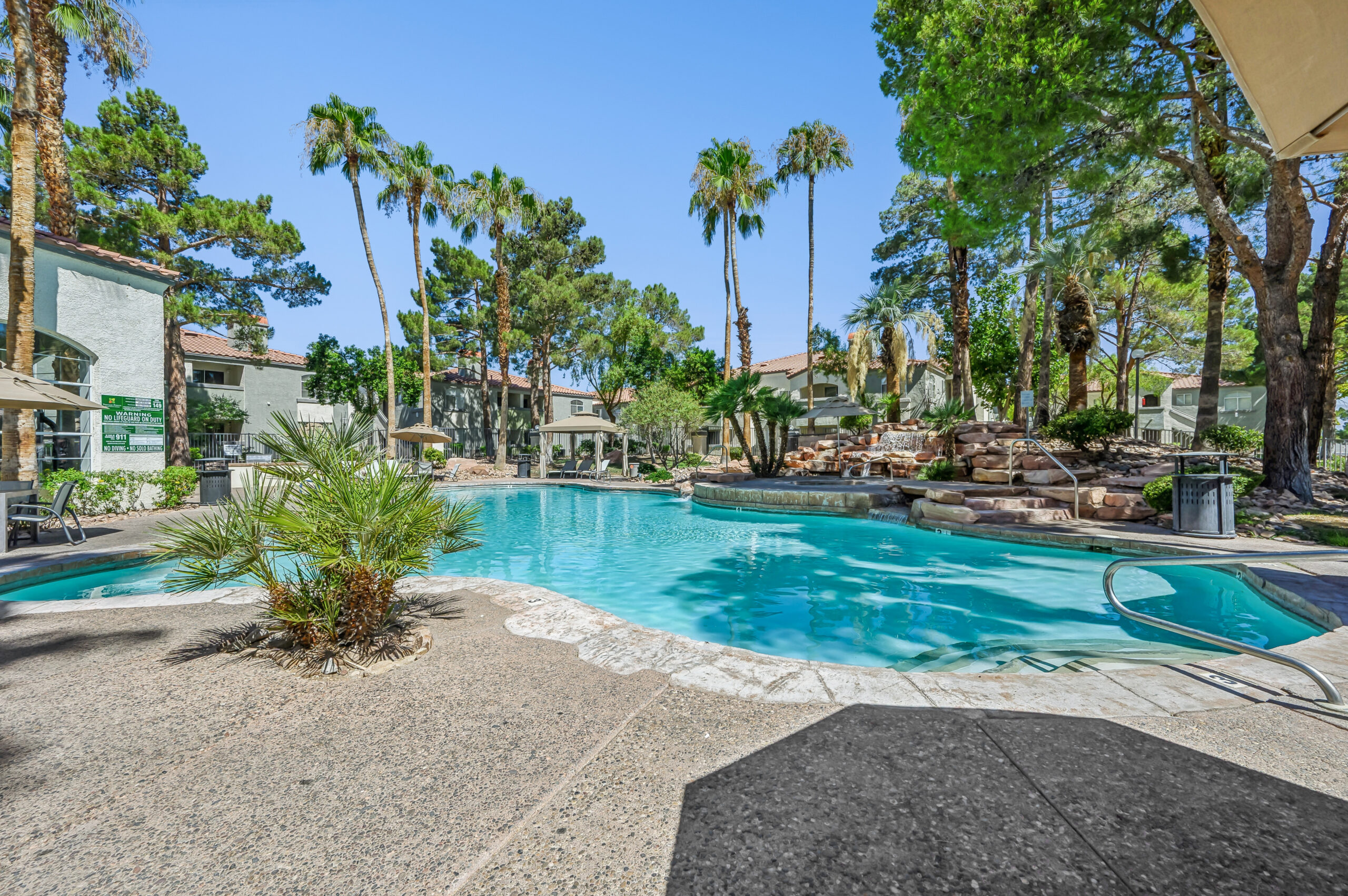 Outdoor swimming pool at Vintage Pointe surrounded by palm trees, mature landscaping, and a rock waterfall.