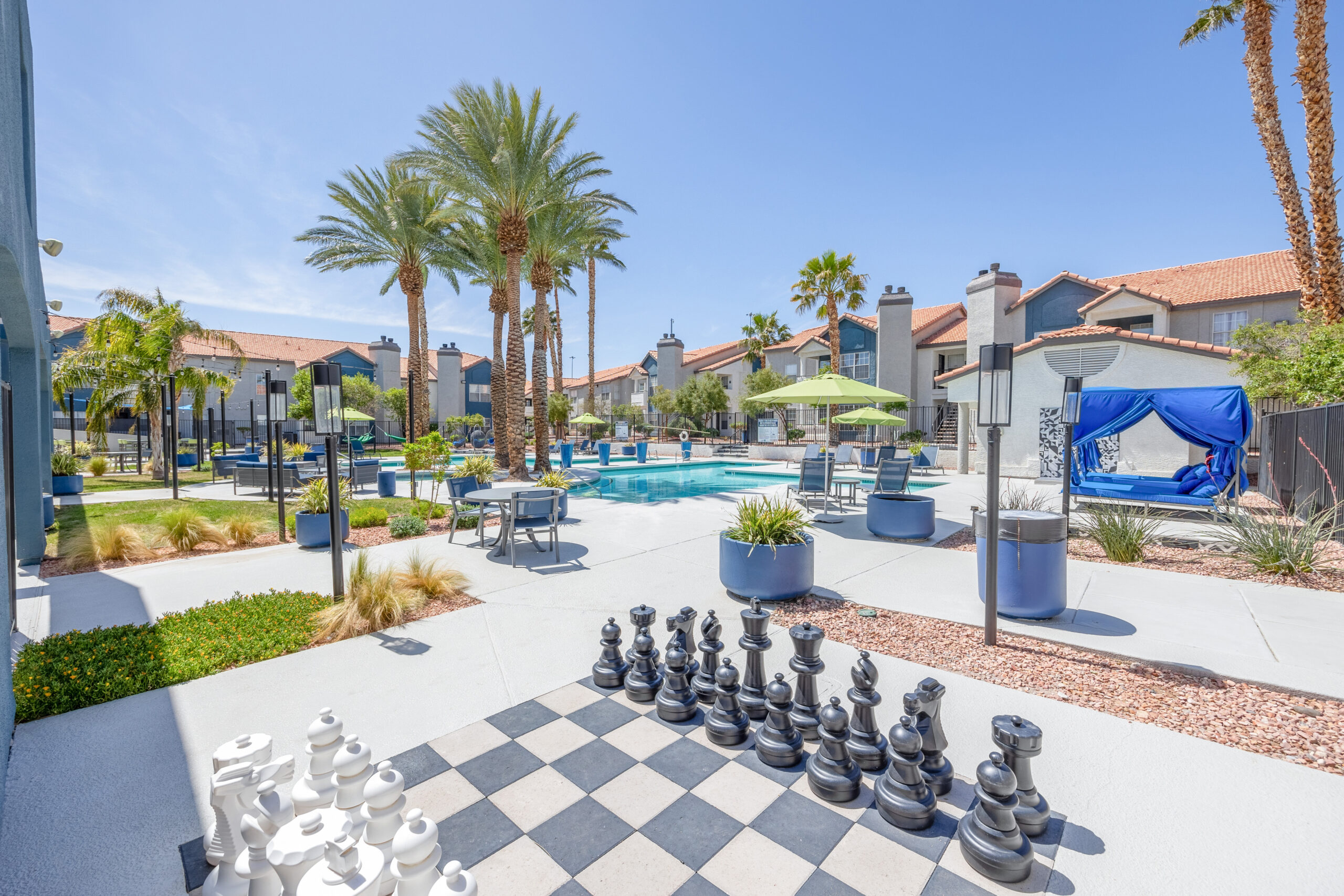 Large outdoor chess board, beautifully landscaped pool courtyard in the background with palm trees, lounge chairs and umbrellas.