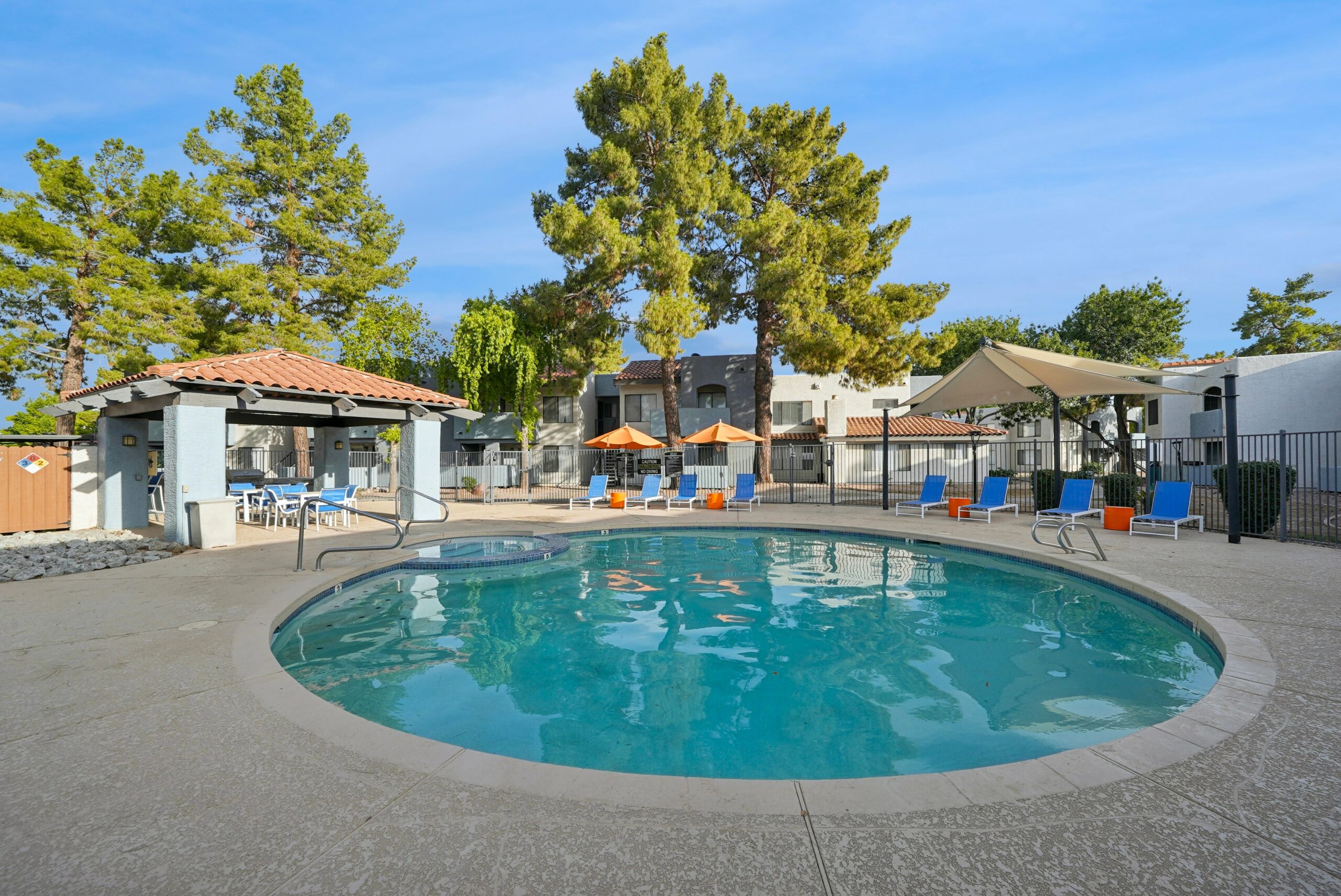 Resort-style swimming pool with a spa, blue lounge chairs, umbrellas, shaded seating, and surrounding apartment homes.