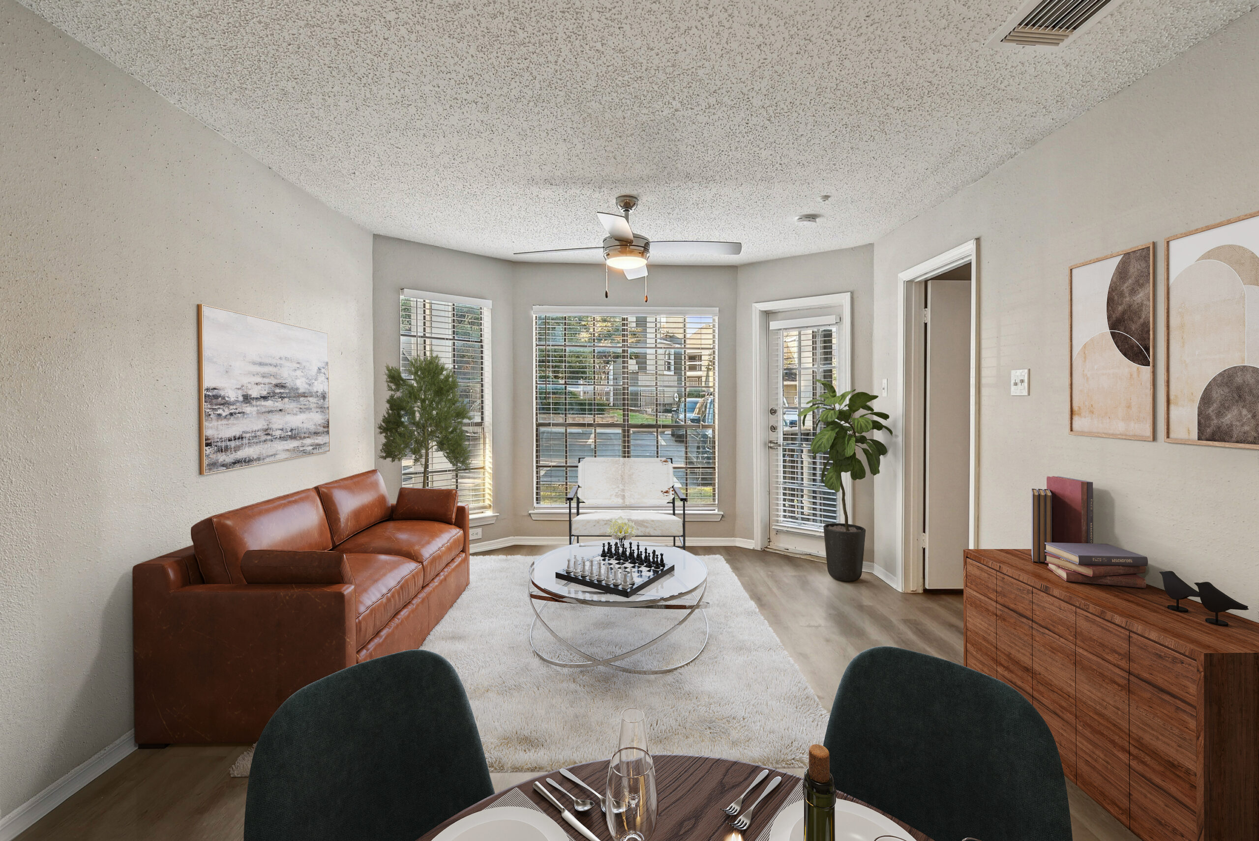 Bright living room apartment in Garland Texas featuring large bay windows, modern staging, wood-style floors, and a leather sofa.