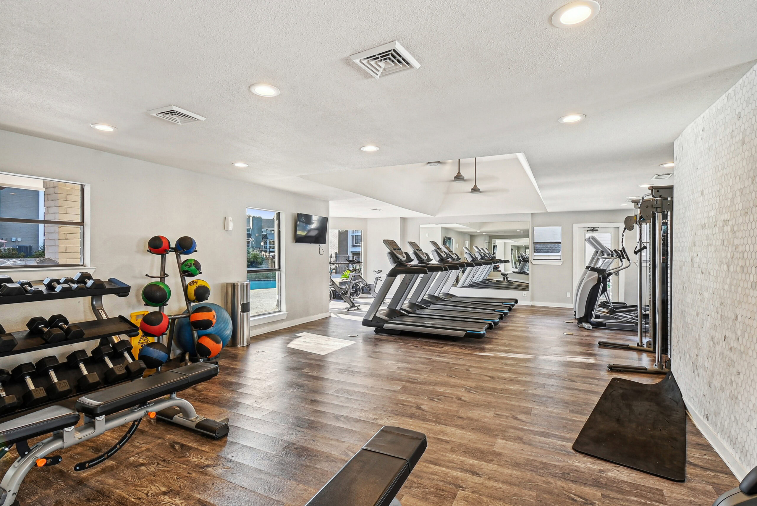 A fully equipped fitness center in the Arbors of Las Colinas apartment building in Garland Texas, with weights, weight balls and treadmills lined against windows