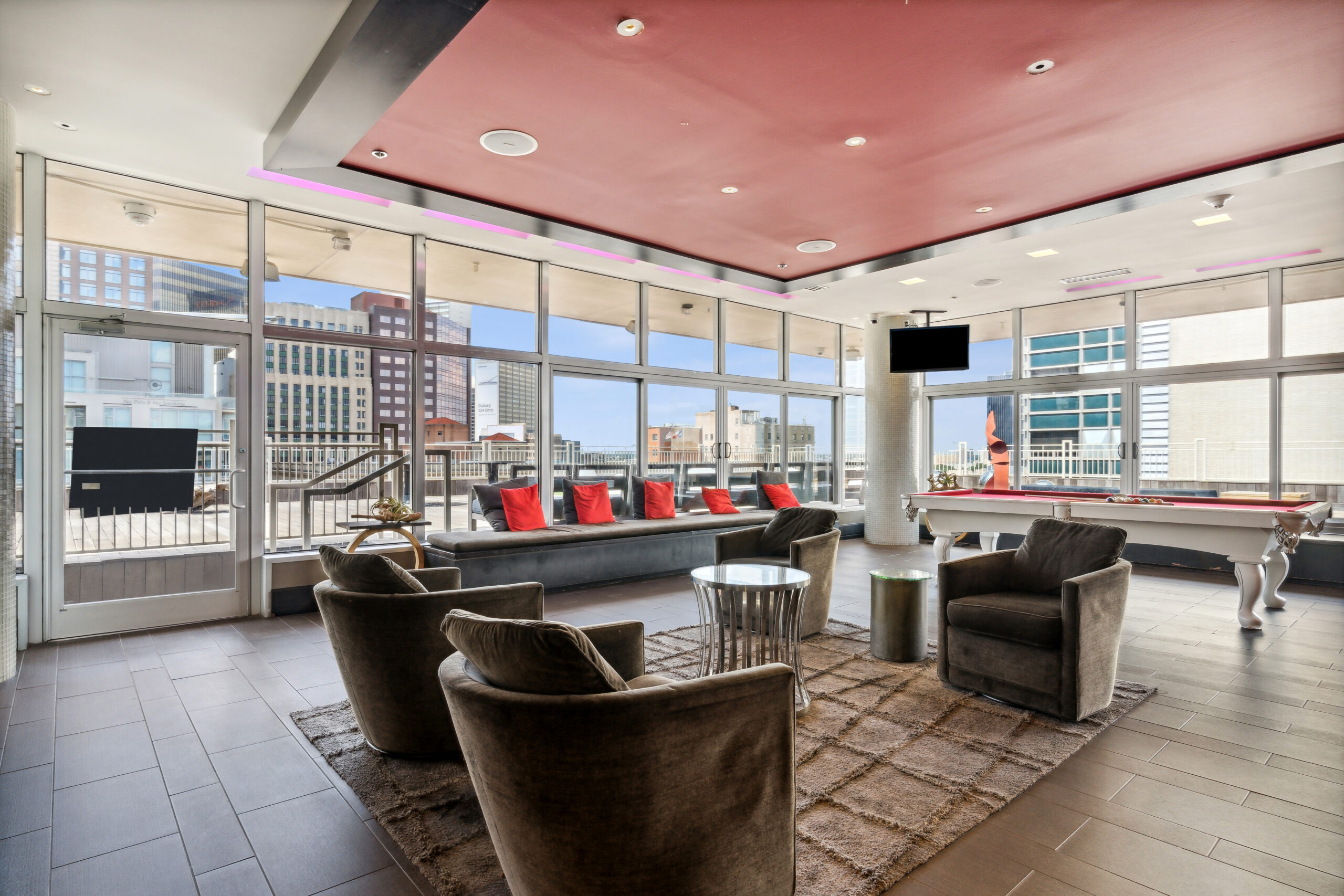 A modern rooftop lounge at The Continentals apartments with suede furniture, a fur rug, red accent throw pillows and window walls offering city views of Dallas, Texas