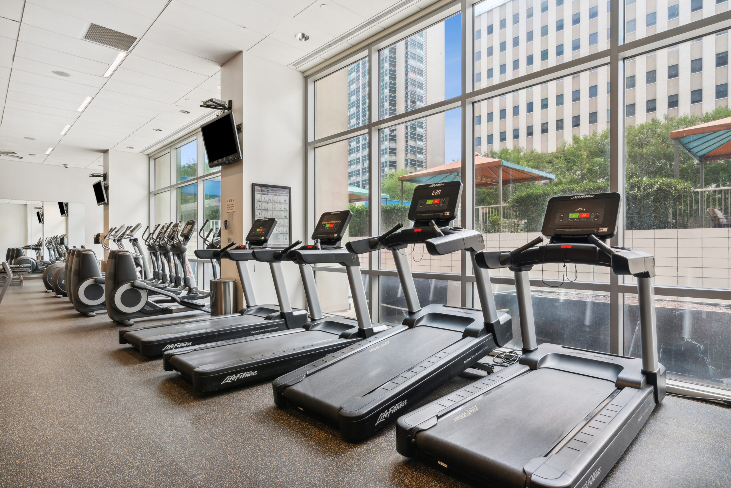 A fitness center with a row of treadmills and gym machines facing a window wall at The Element luxury apartments in Dallas Texas