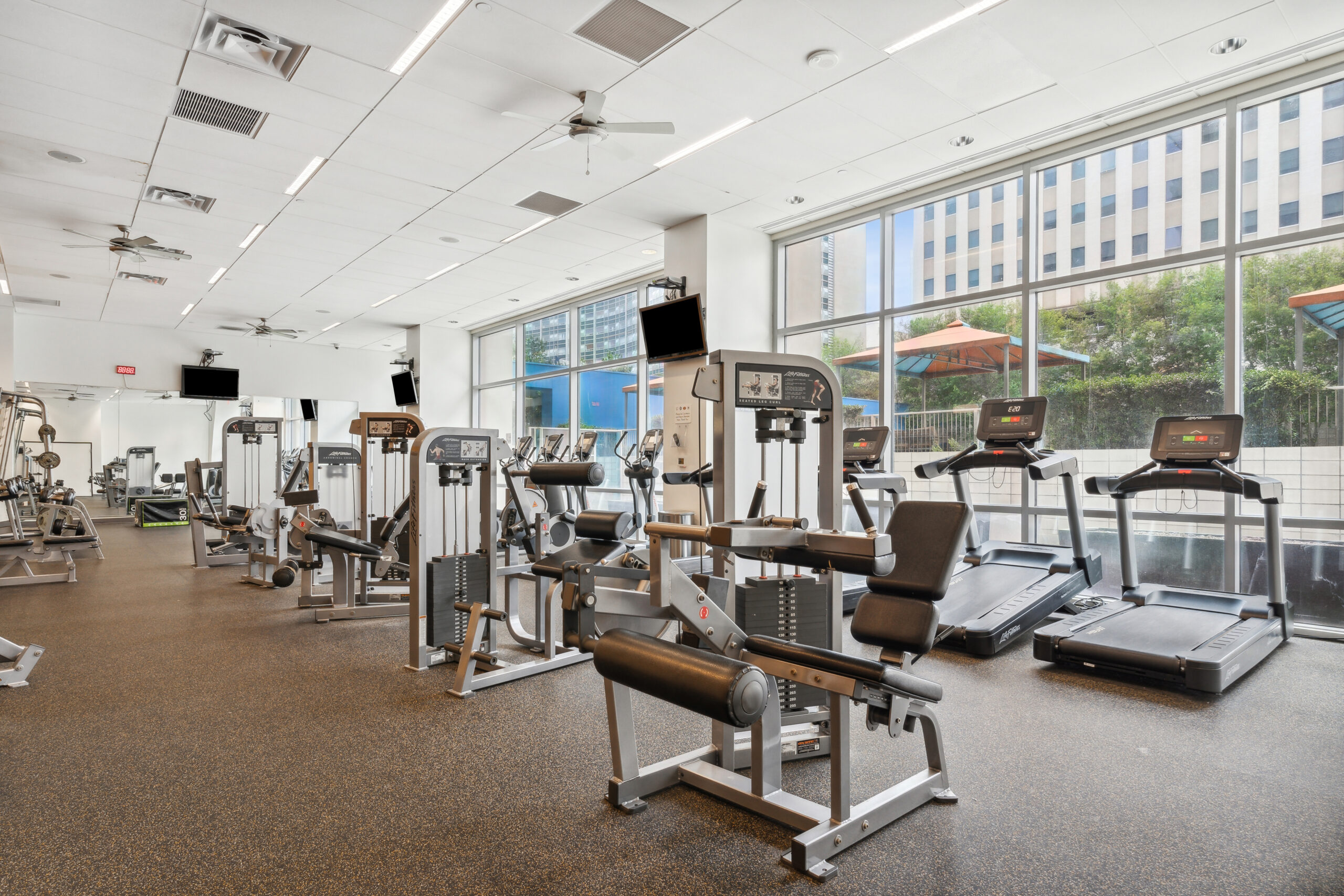 A fully equipped fitness center with several weight and cardio machines, and natural lighting from the window wall at The Element luxury apartments in Dallas Texas