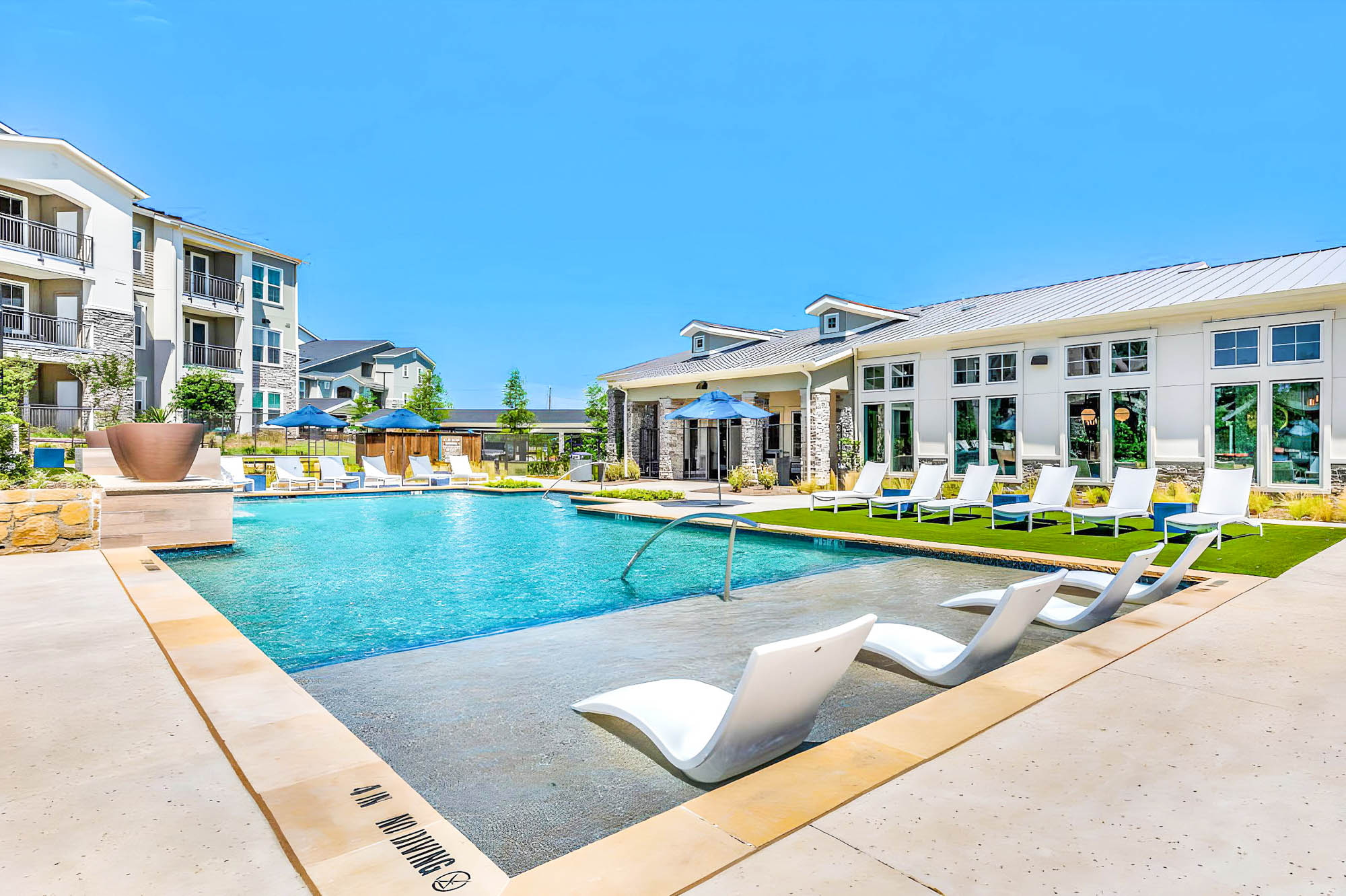 A modern pool with lounging chairs on a step in the pool and on a lawn, at the Embree Hill apartments in Garland, Texas