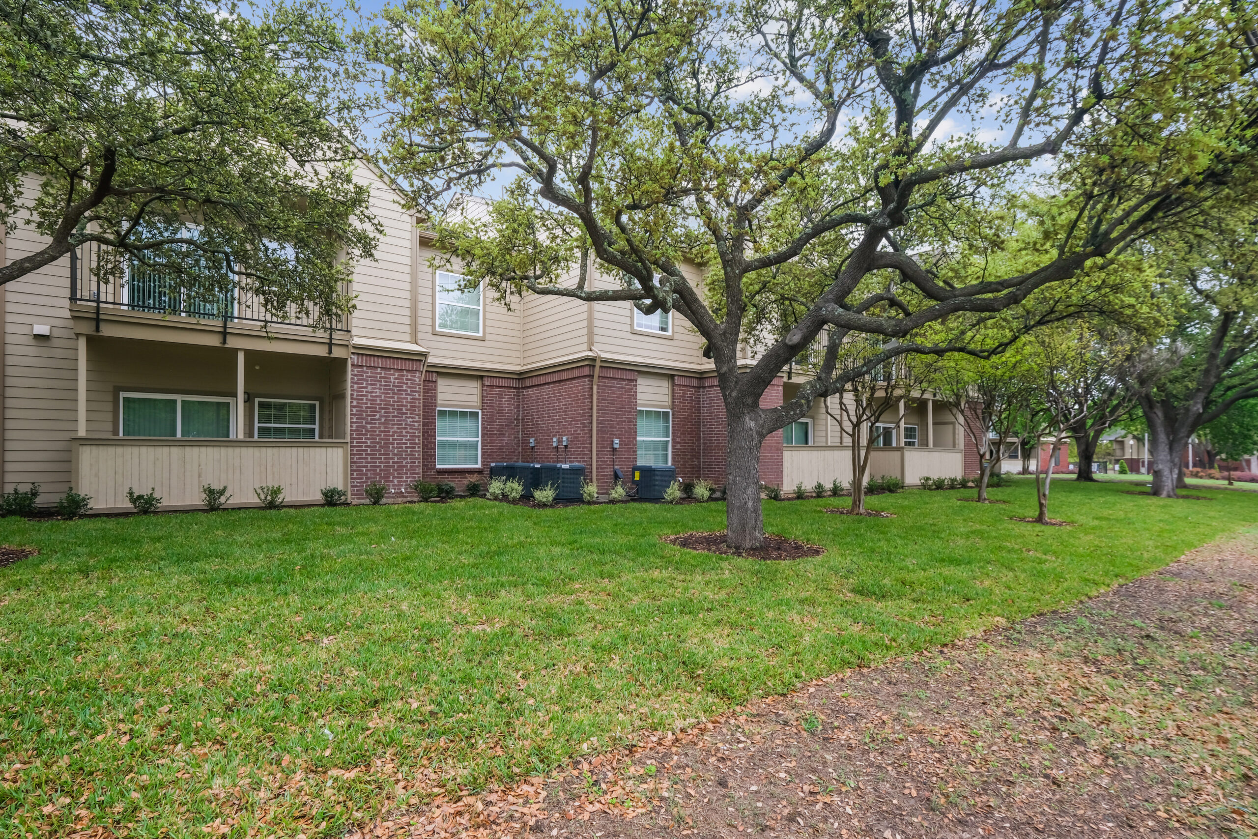 Scenic exterior view of the Gables of Mckinney apartments in Mckinney Texas featuring spacious green lawns, mature trees, and brick-and-siding architecture.