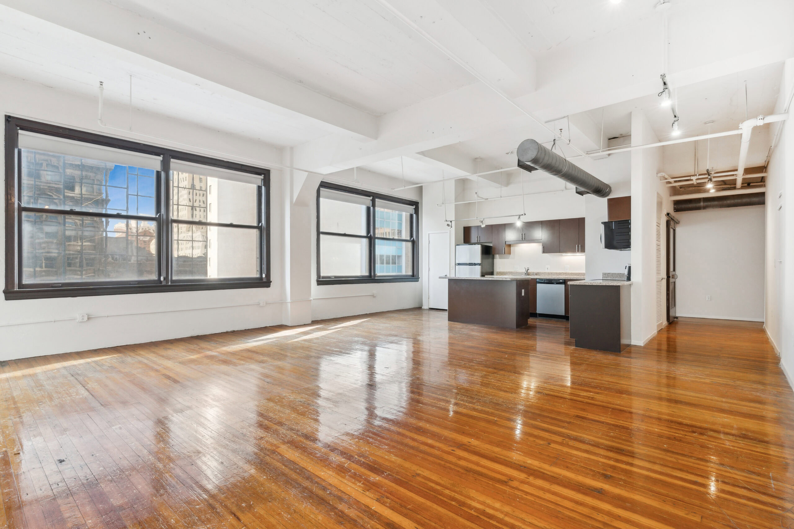 Open plan living and dining room area with large windows, polished wood flooring and exposed piping in a vacant boutique apartment at The Wilson in Dallas Texas