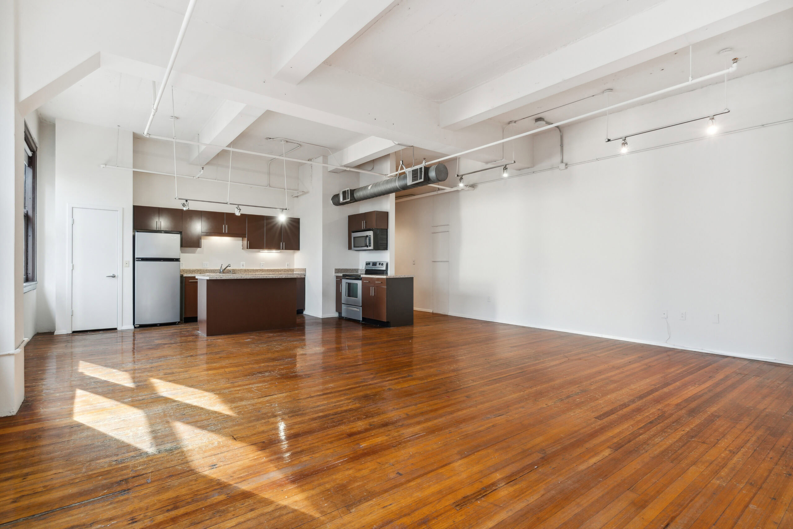 Open plan living and dining room area with natural lighting, polished wood flooring and exposed piping in a vacant boutique apartment at The Wilson in Dallas Texas