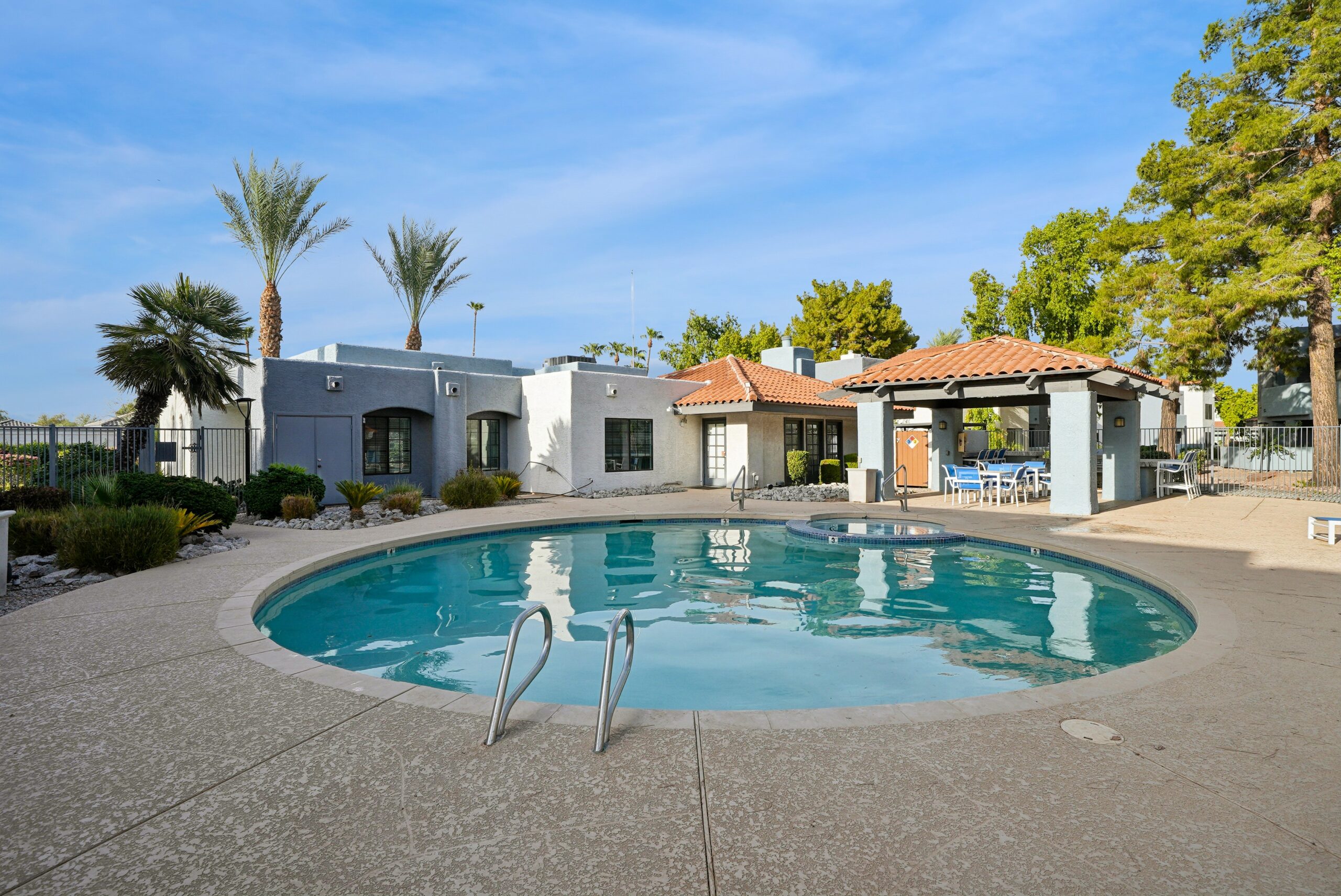 Outdoor swimming pool and spa with covered poolside seating, palm trees, and nearby community buildings.